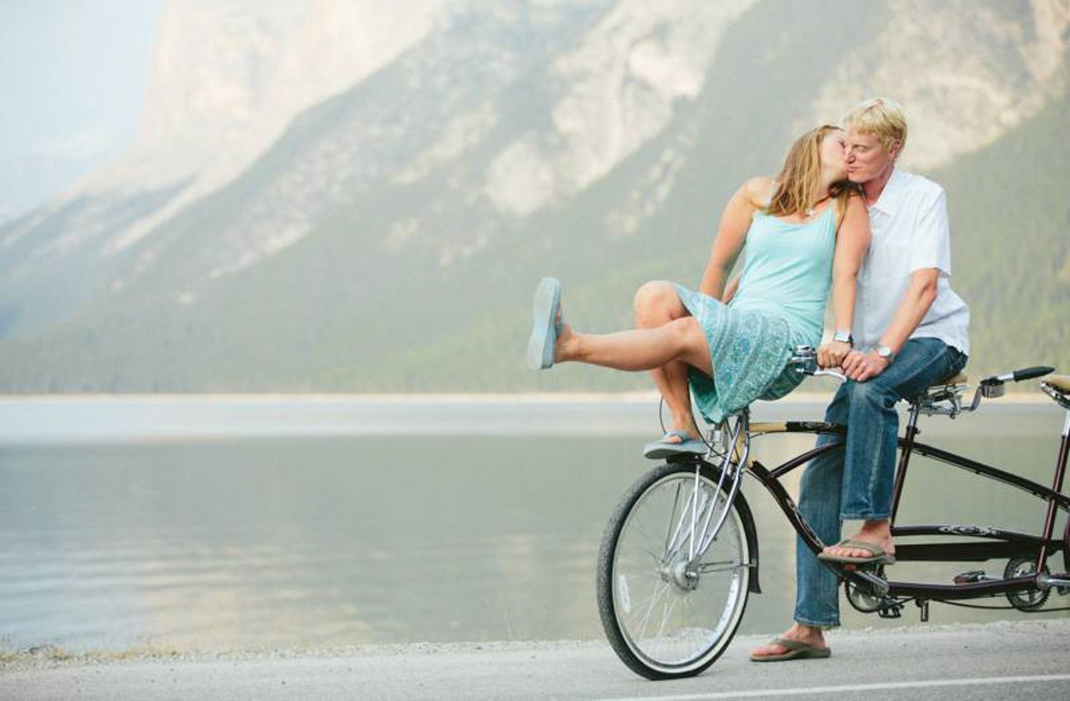 A cyclist performs a daring bike trick sequence on a mountain trail with scenic lake and mountain views in the background at lake Minnewanka