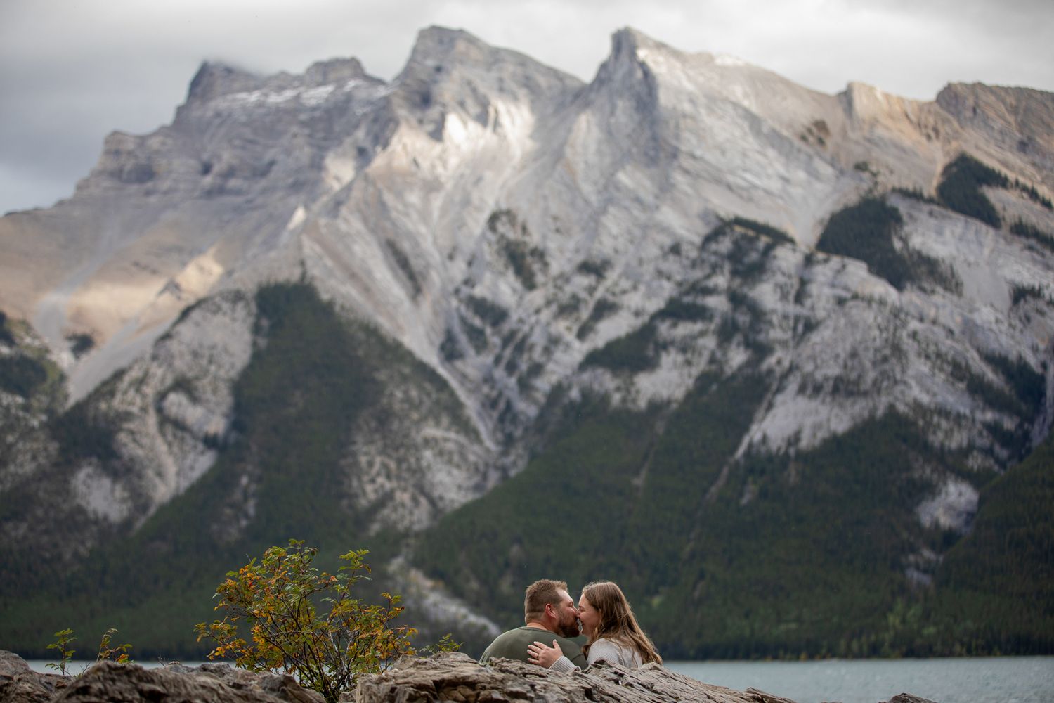 A couple shares a romantic moment on a rocky overlook with majestic Banff mountains and Lake Minnewanka in the background.