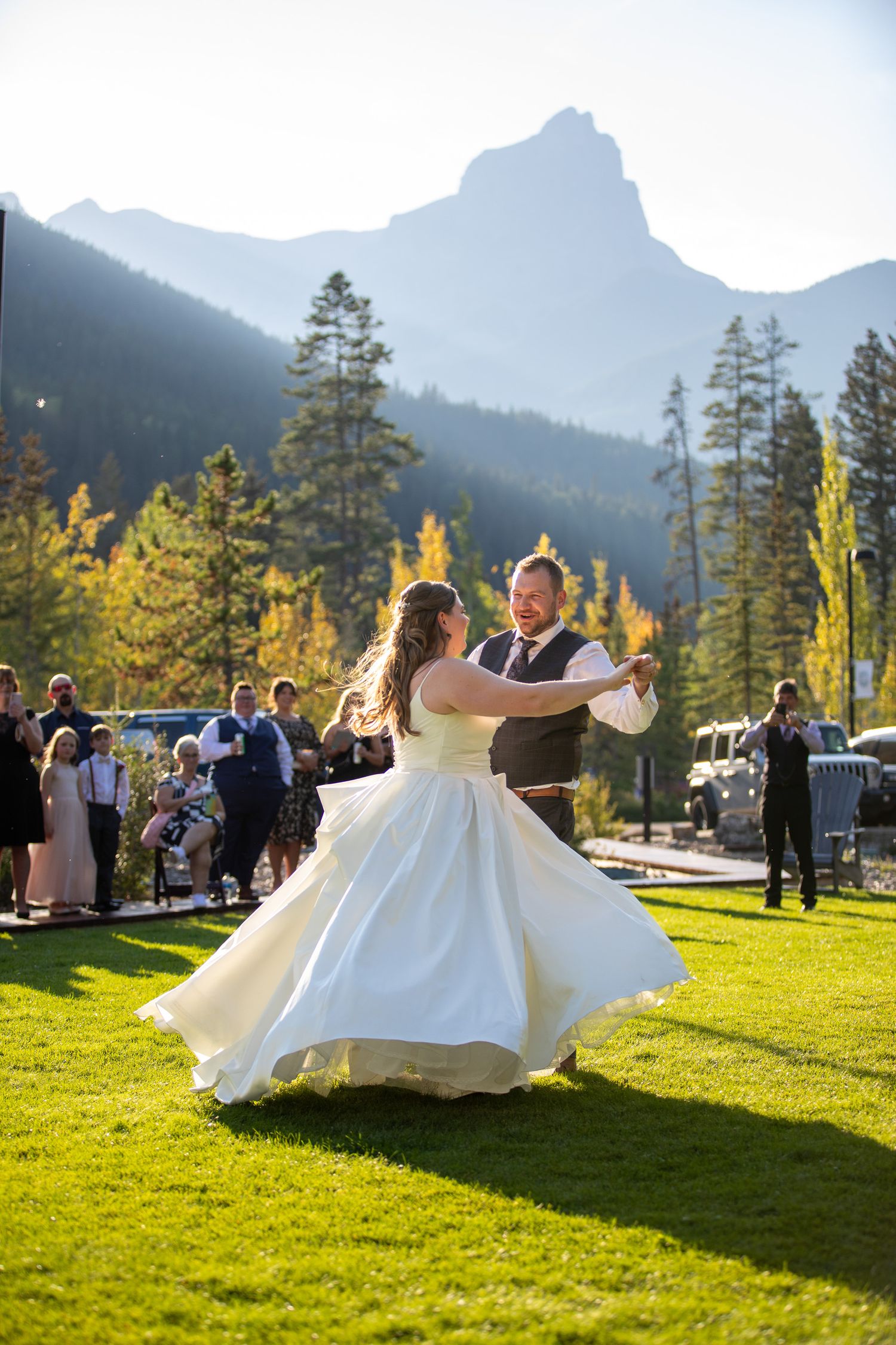 A couple spins joyfully on a grassy lawn with mountain peaks in the background during a wedding celebration at Stewart Creek, Canmore, AB