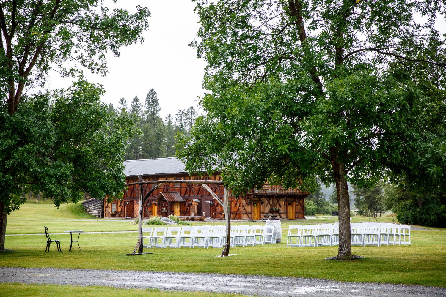 Rustic wooden barn wedding venue at Cherry creek estate with white chairs arranged outdoors on green lawn surrounded by tall trees.