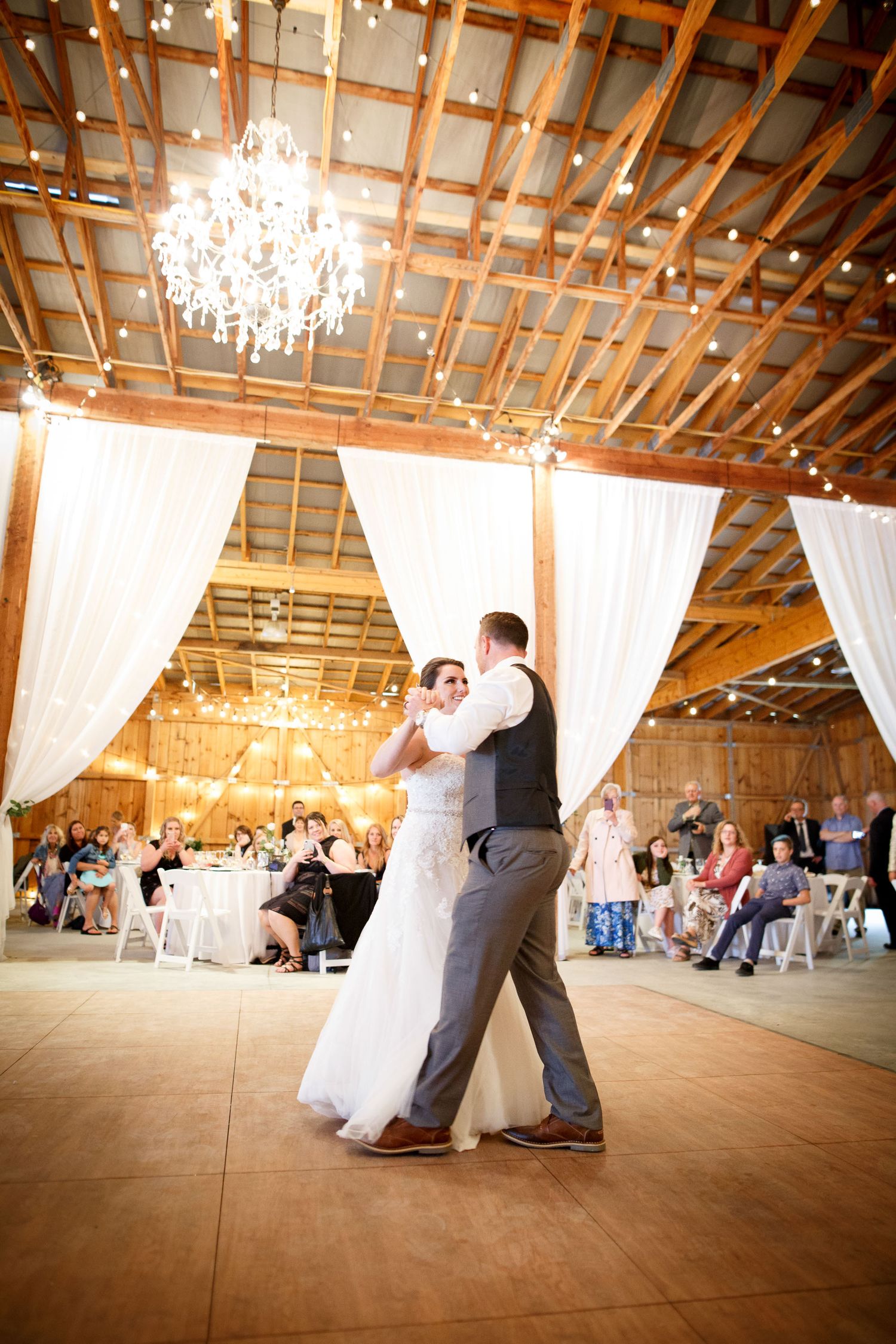 Bride and groom share their first dance in a rustic barn wedding venue with chandelier and string lights at Cherry creek estate