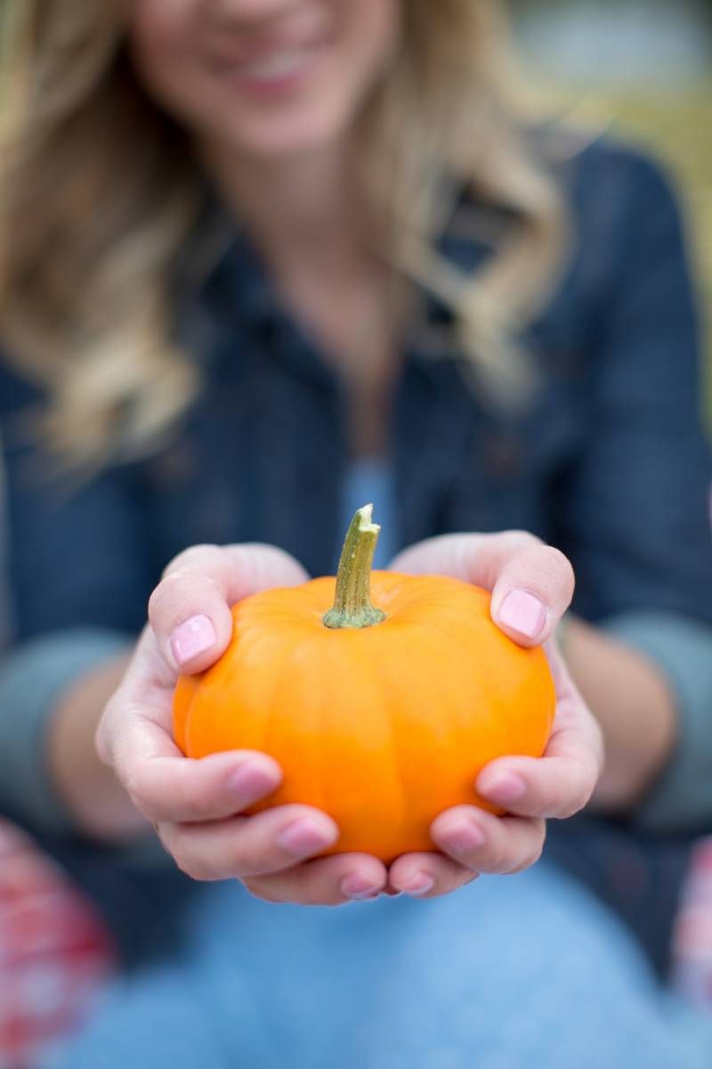 Woman holding a small orange pumpkin toward camera in autumn setting at Fish Creek Park in Calgary.