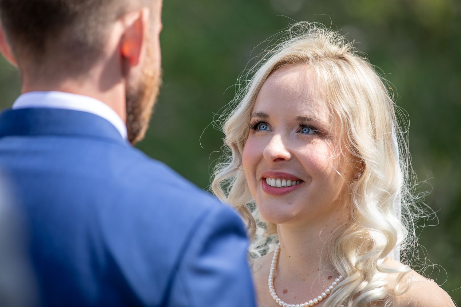 Smiling blonde bride in pearl necklace gazes lovingly at groom in blue suit during outdoor wedding ceremony at Silvertip gazebo in Canmore