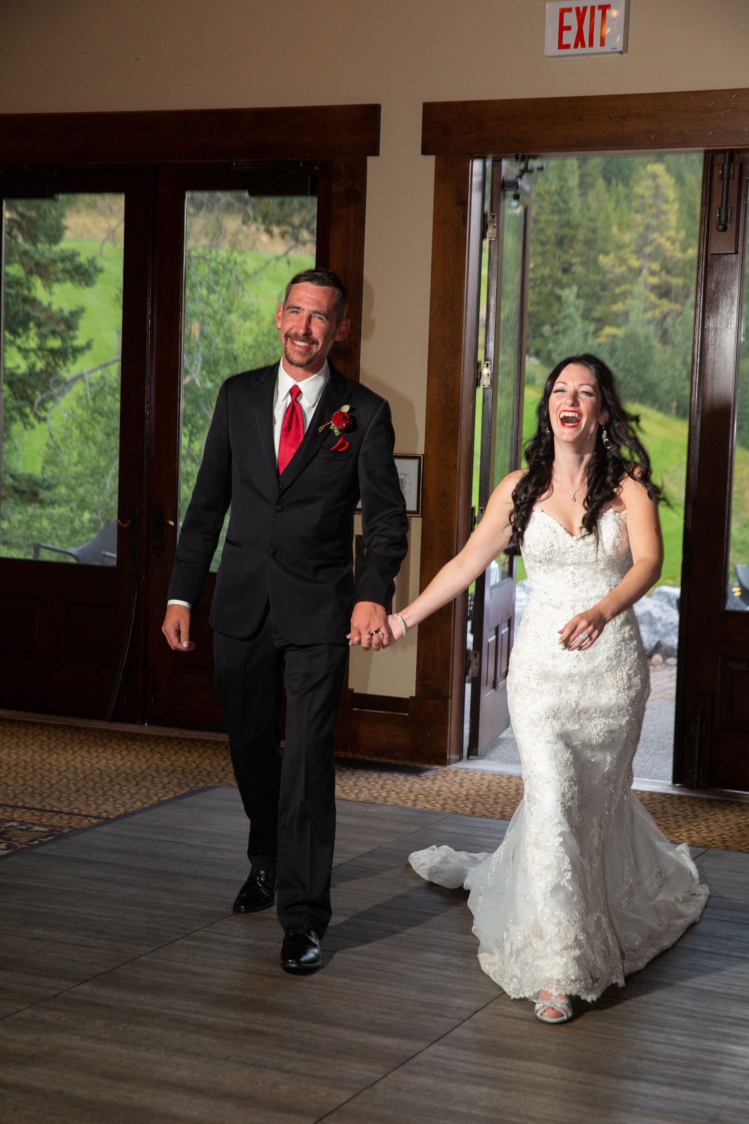Bride and groom holding hands smiling as they enter their wedding reception, bride in lace gown, groom in black suit with red tie.