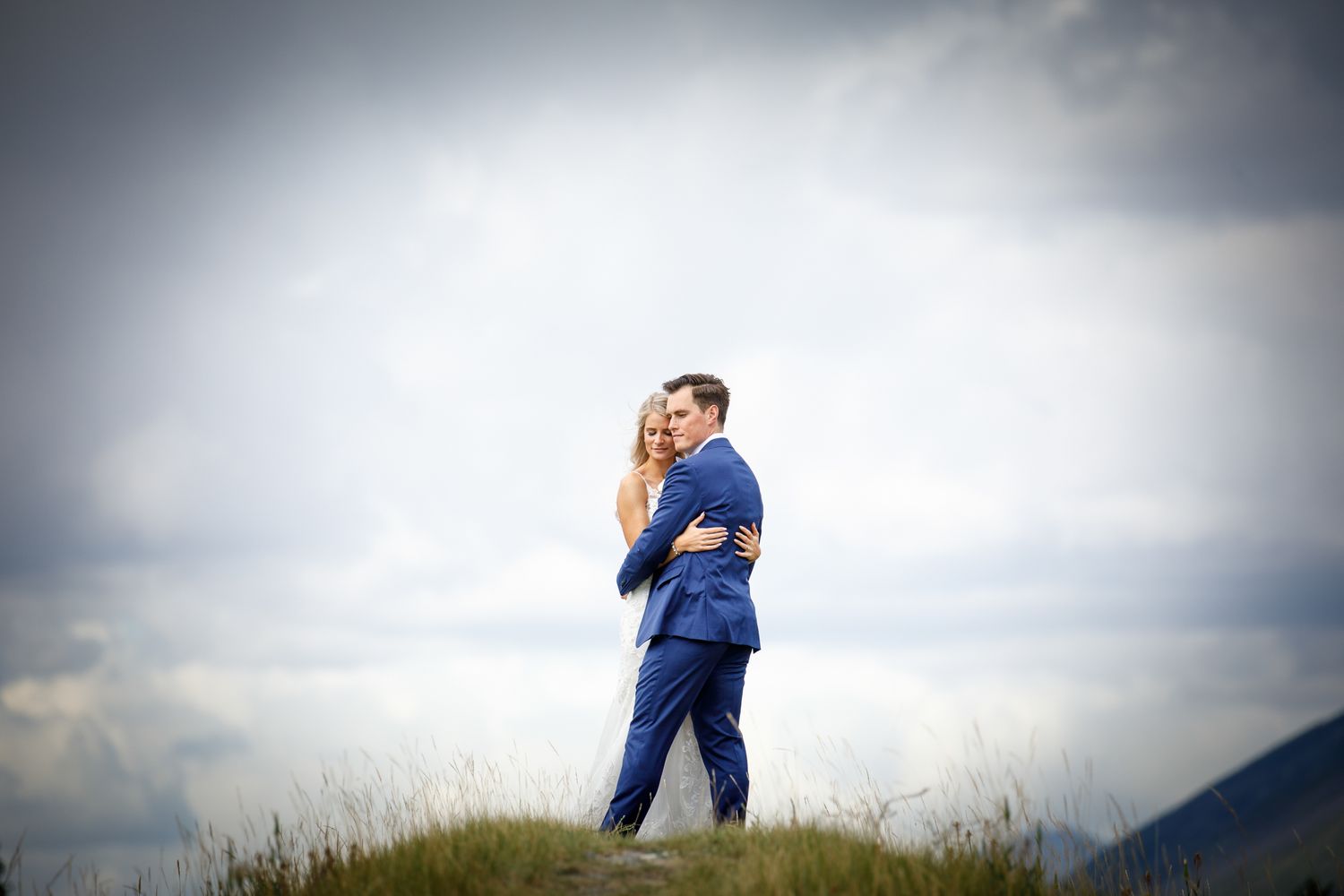 A couple in navy blue outfits embrace on a hilltop against a dramatic cloudy sky at sunset Canmore Three Sisters in Canmore, AB.