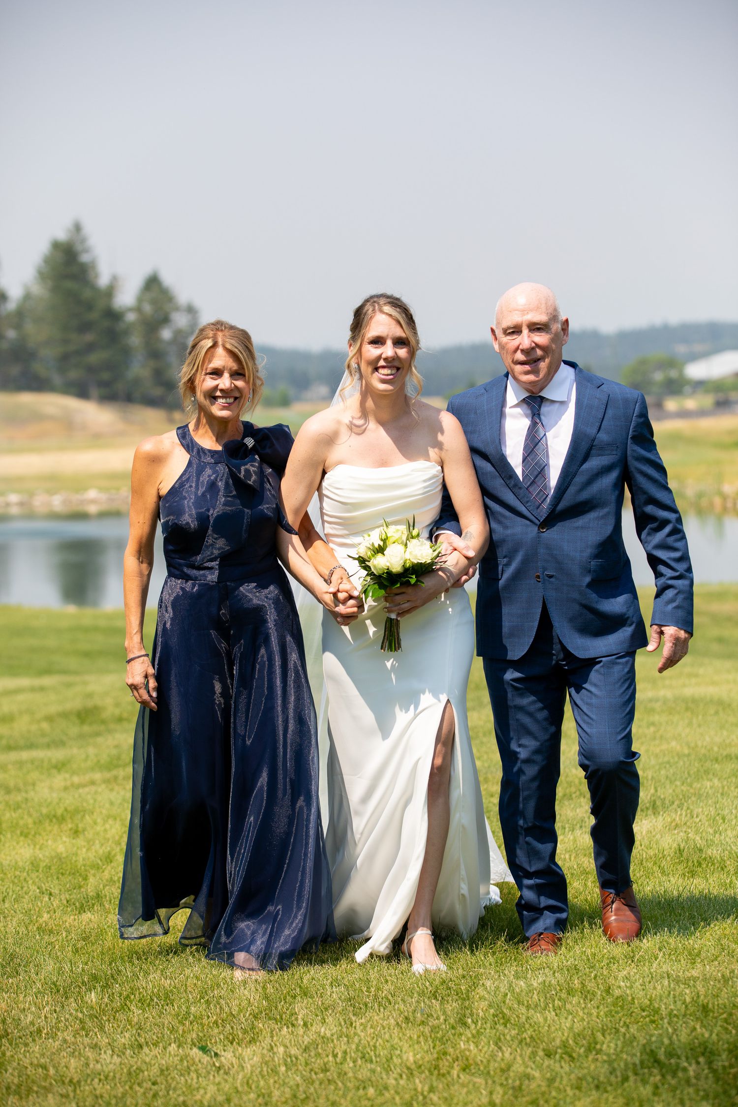Bride in white gown with bouquet walks with parents in navy attire on green lawn near lake at Swansea Ranch in Invermere, BC