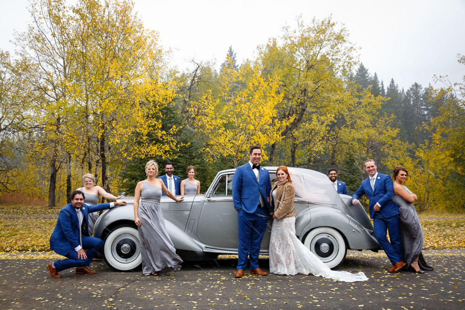 A wedding party poses with a vintage silver car in a scenic autumn setting with golden foliage in the background. Canmore Alberta.