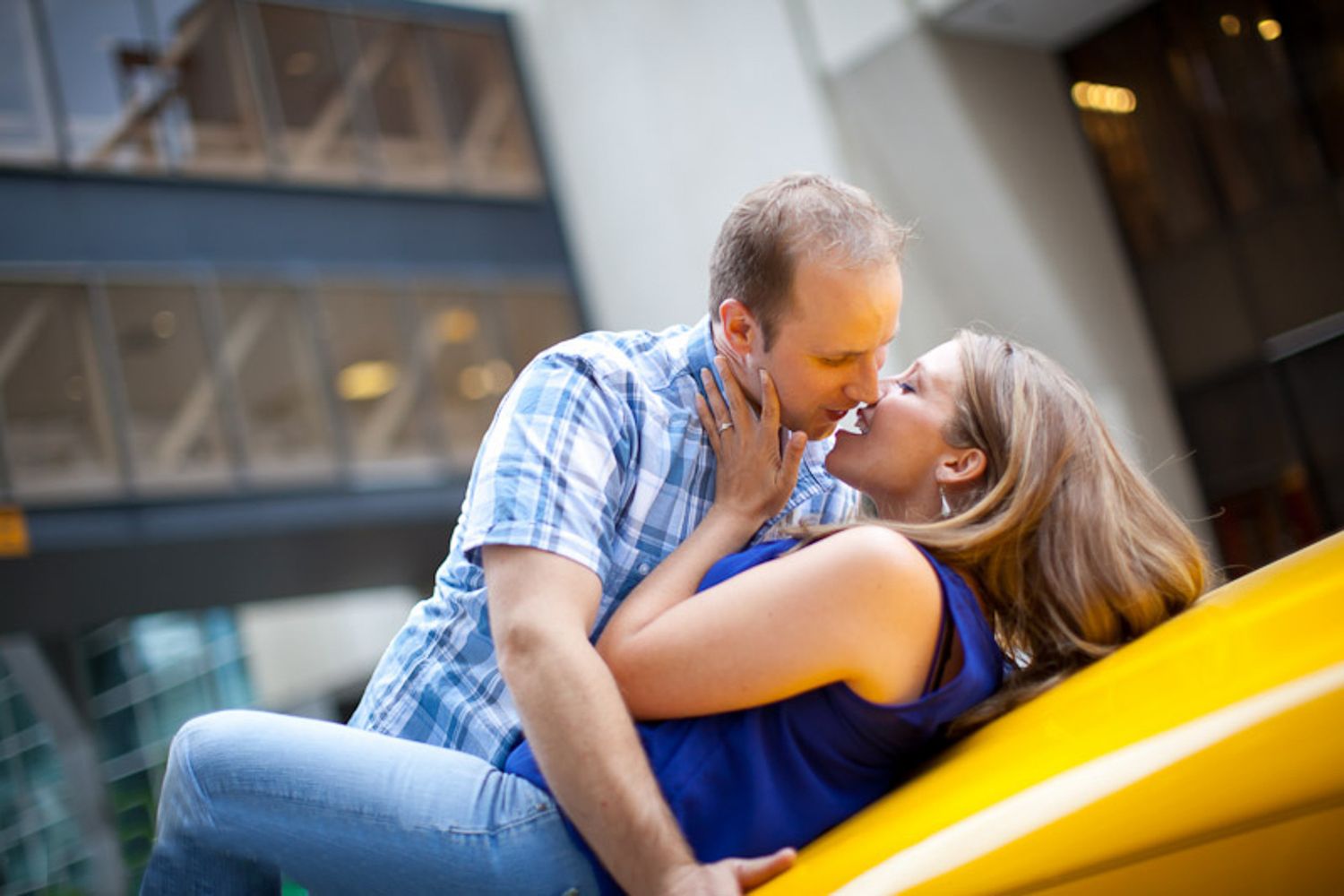 A couple shares a romantic kiss while leaning against a bright yellow taxi cab in an urban Calgary downtown, AB