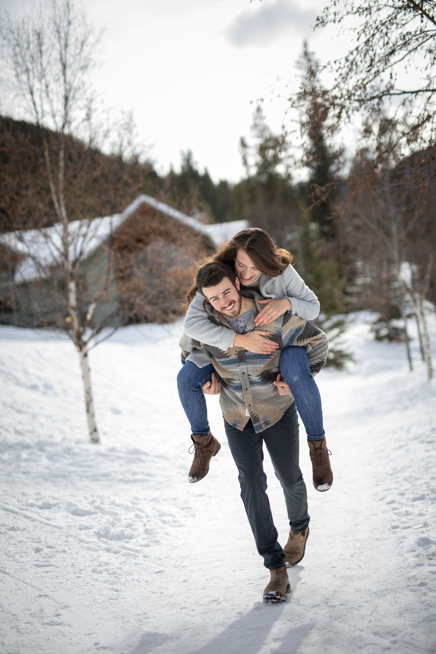 A couple shares a playful piggyback ride moment on a snowy winter path with a cabin in the background at Grey Wolf Panorama, BC