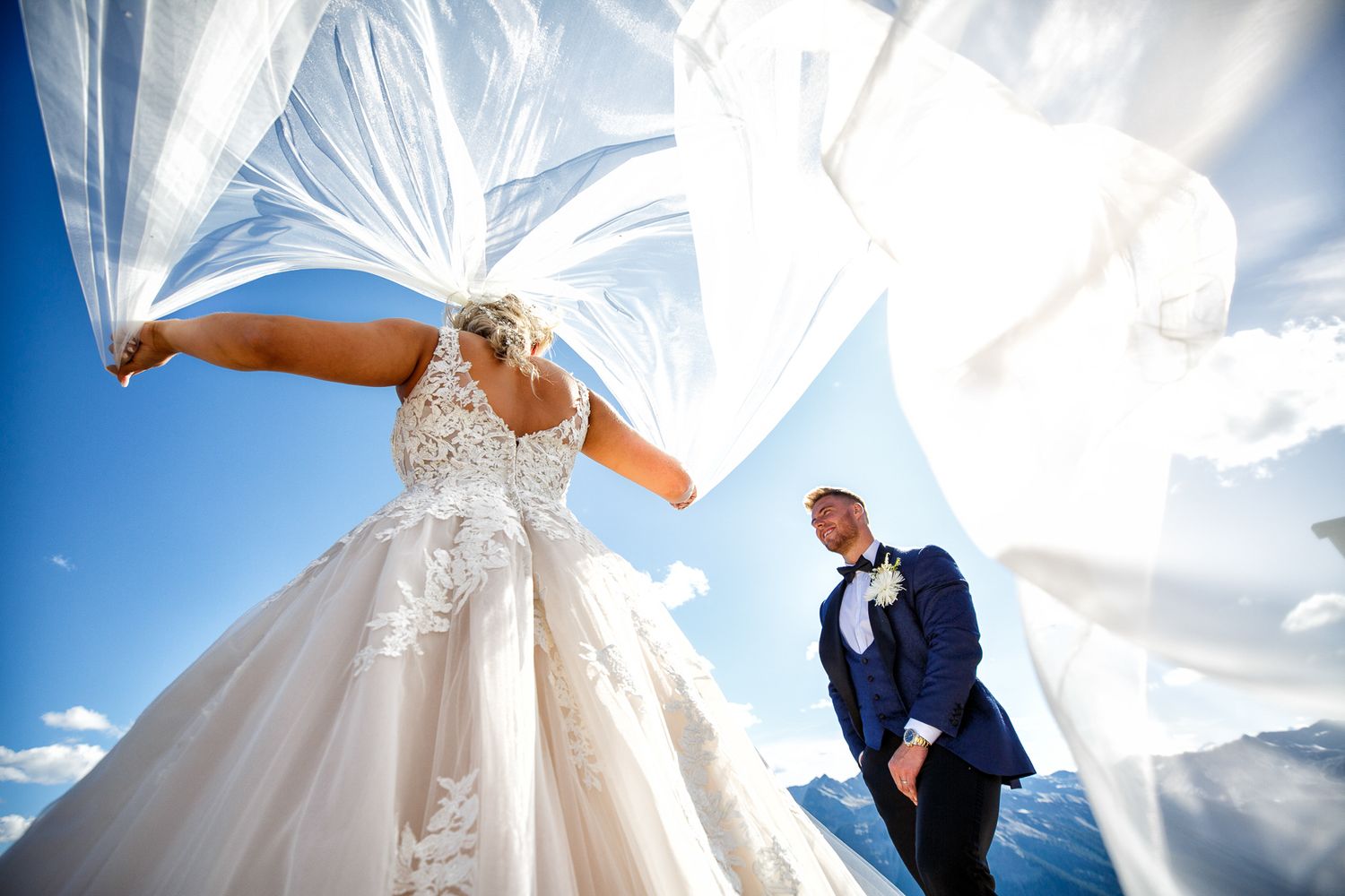 White wedding dress billowing dramatically in the wind against a bright blue sky with clouds. Kicking Horse resort, Golden, BC.