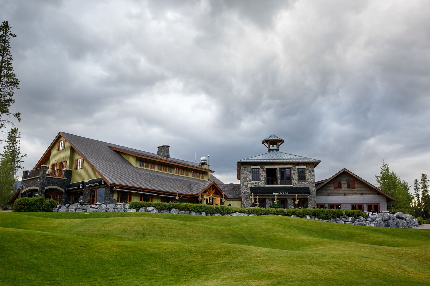 A panoramic view of a golf course clubhouse with rolling hills and cloudy skies in the background. Silvertip resort in Canmore.