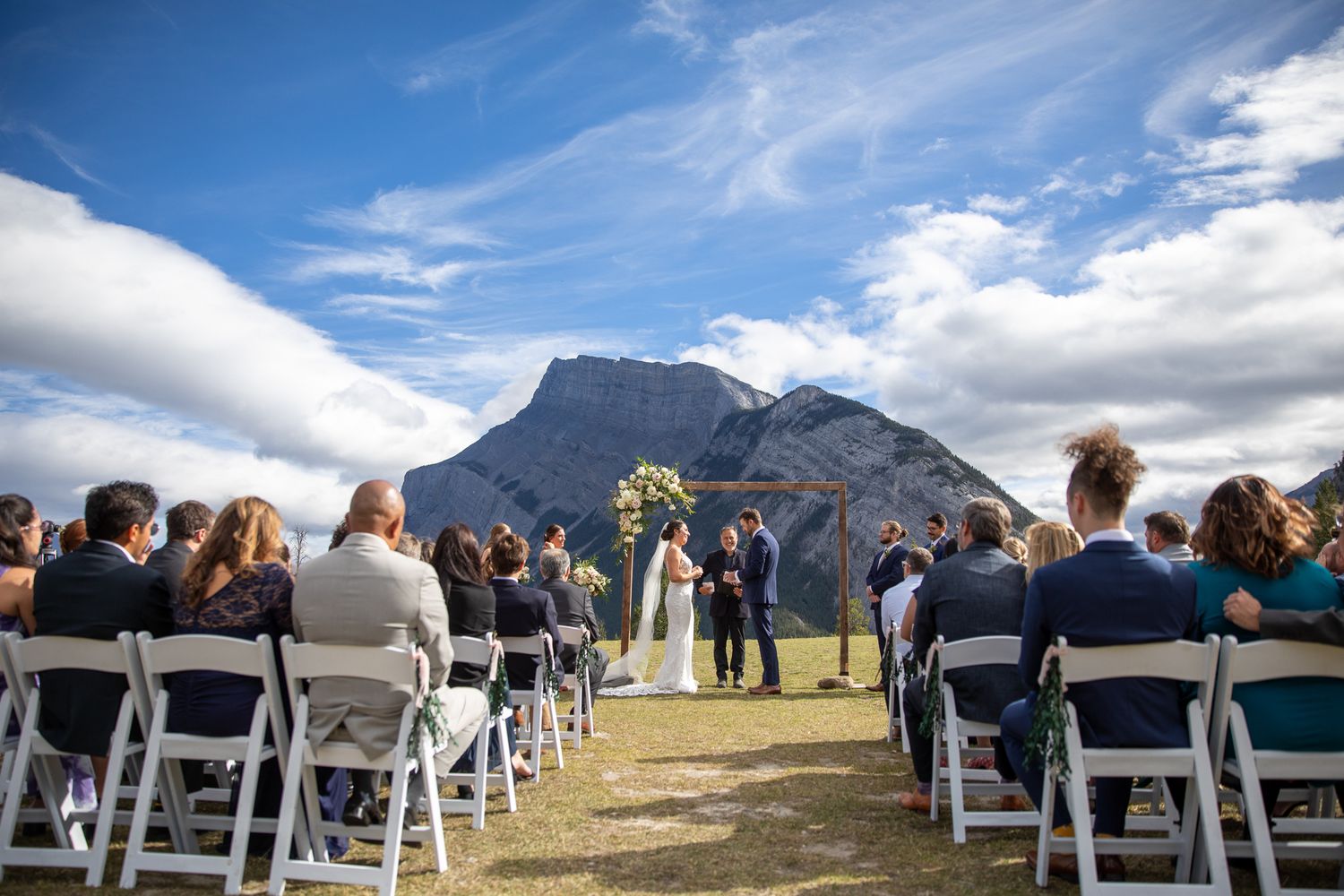Wedding ceremony guests seated in white chairs face a majestic mountain backdrop under bright blue skies. Tunnel Mountain Reservoir Banff.