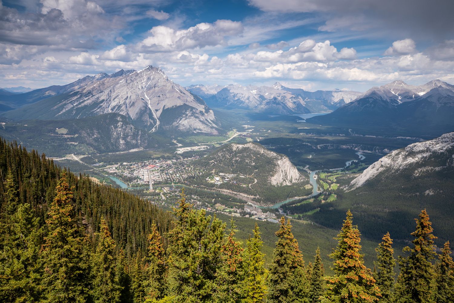 Aerial view of Banff townsite nestled in a mountain valley surrounded by evergreen forests and Rocky Mountains under cloudy skies.