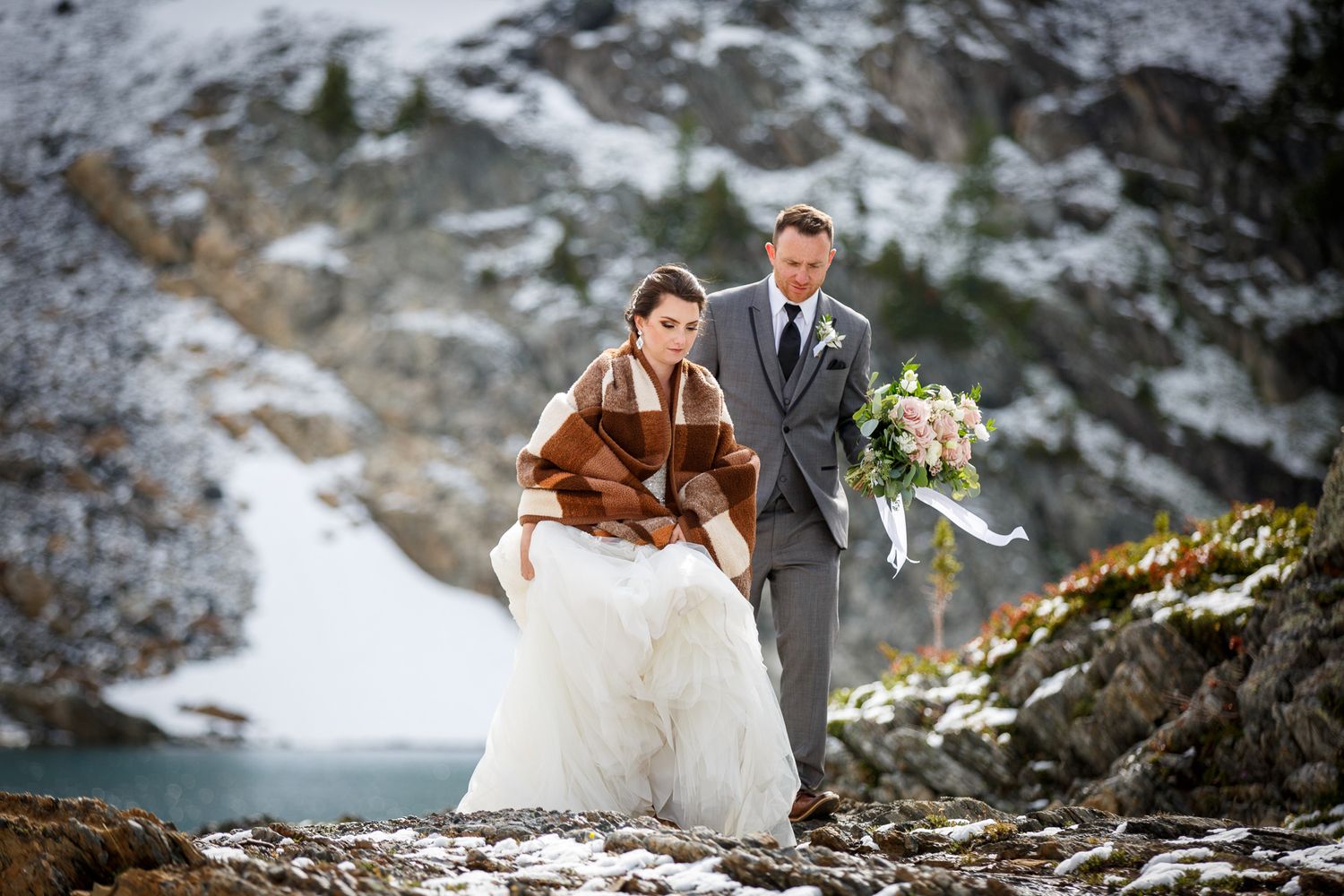 Bride in white gown and brown fur shawl walks with groom in grey suit through snowy mountain landscape in the backcountry of Kimberley, BC