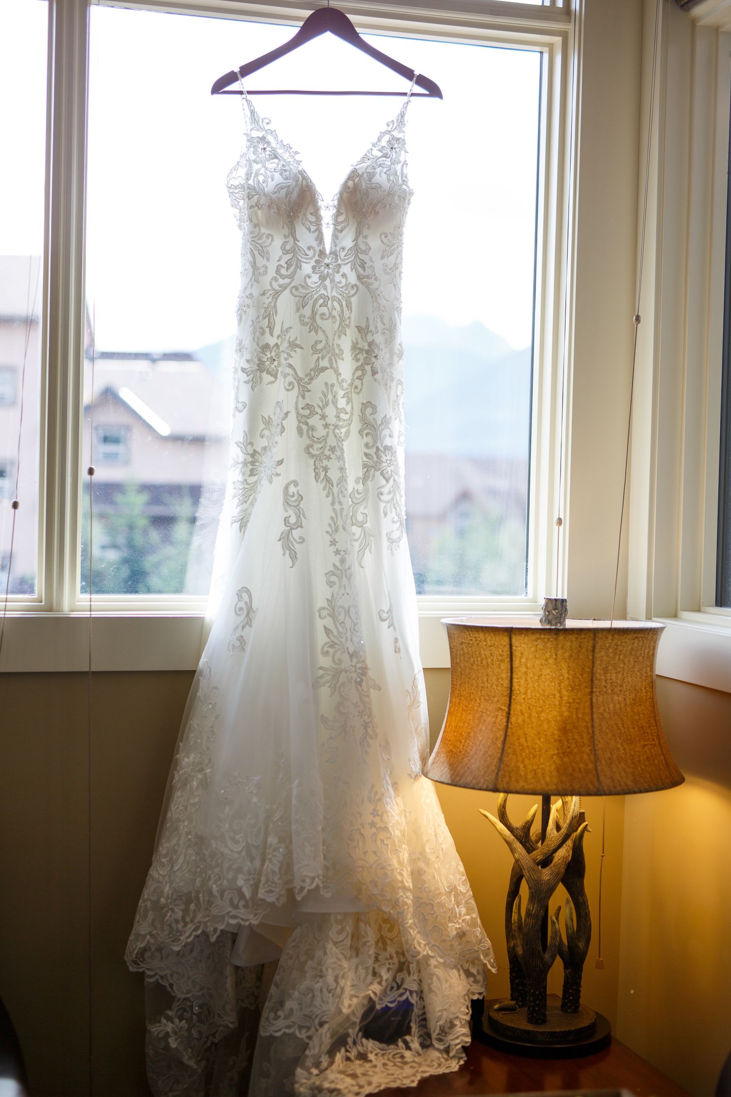 A white lace wedding dress hangs by a window with a warm glowing table lamp in the foreground.