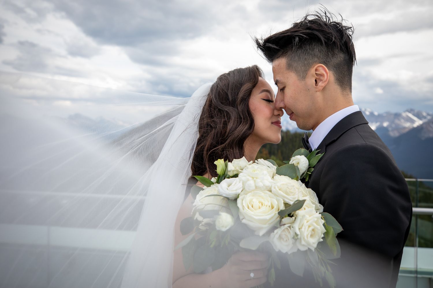Bride and groom sharing a romantic moment with white rose bouquet and veil flowing against mountain backdrop.