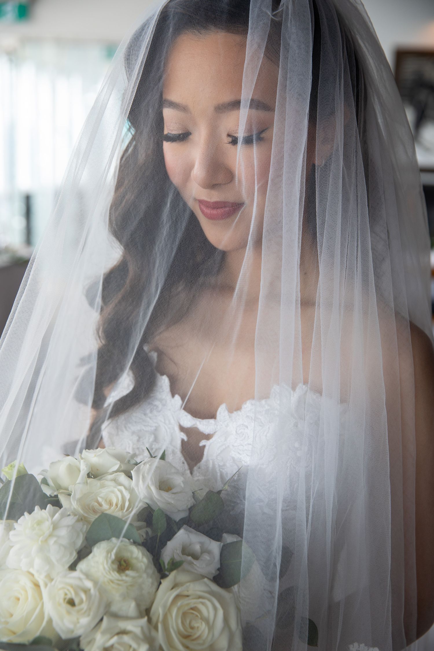 Bride in white lace gown and veil holding white rose bouquet, looking down elegantly.