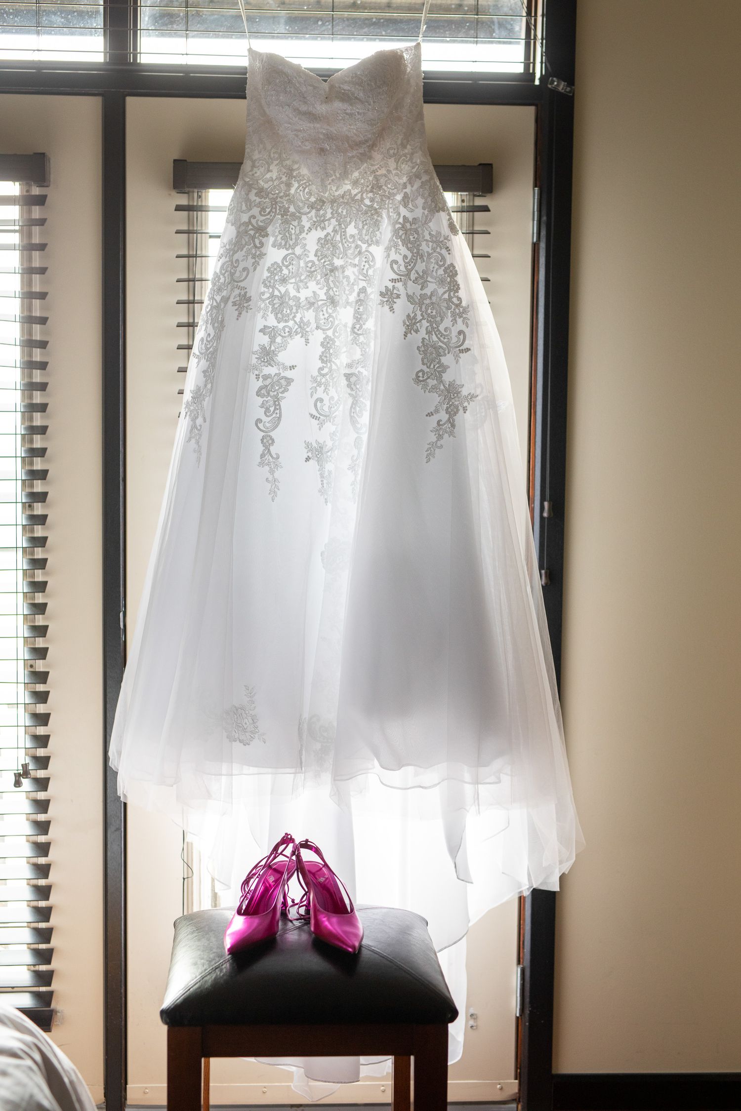 White lace strapless wedding gown hanging by window with pink high heel shoes displayed on chair below. Copperpoint resort,  Invermere.