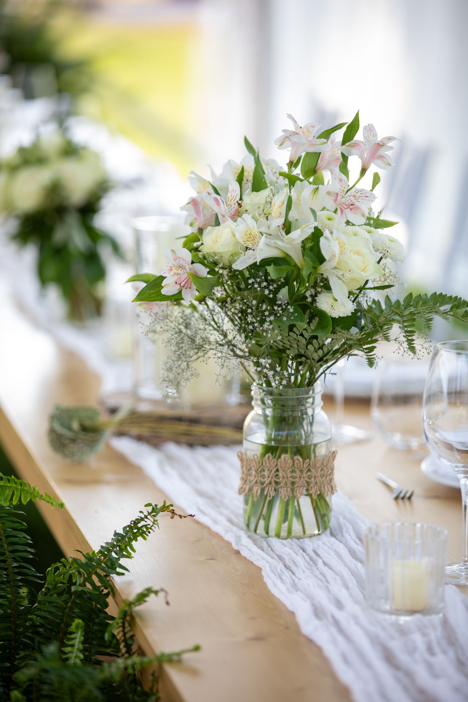 Elegant wedding table center piece with white flowers in a burlap-wrapped mason jar on a lace runner at Swansea Ranch in Invermere, BC