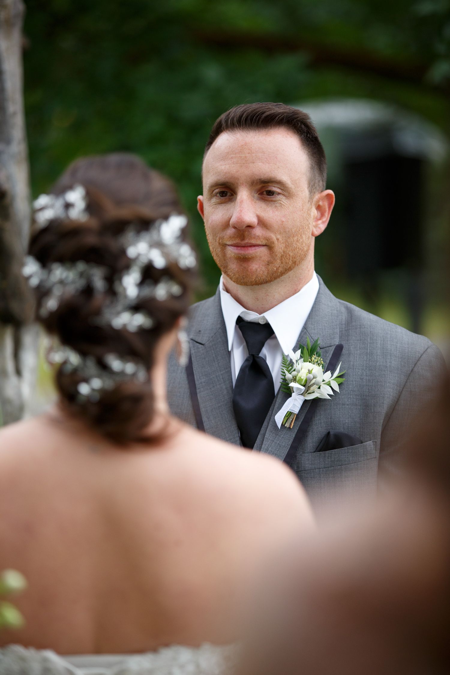Groom in gray suit with boutonniere smiling at bride during outdoor wedding ceremony. Cherry creek estate