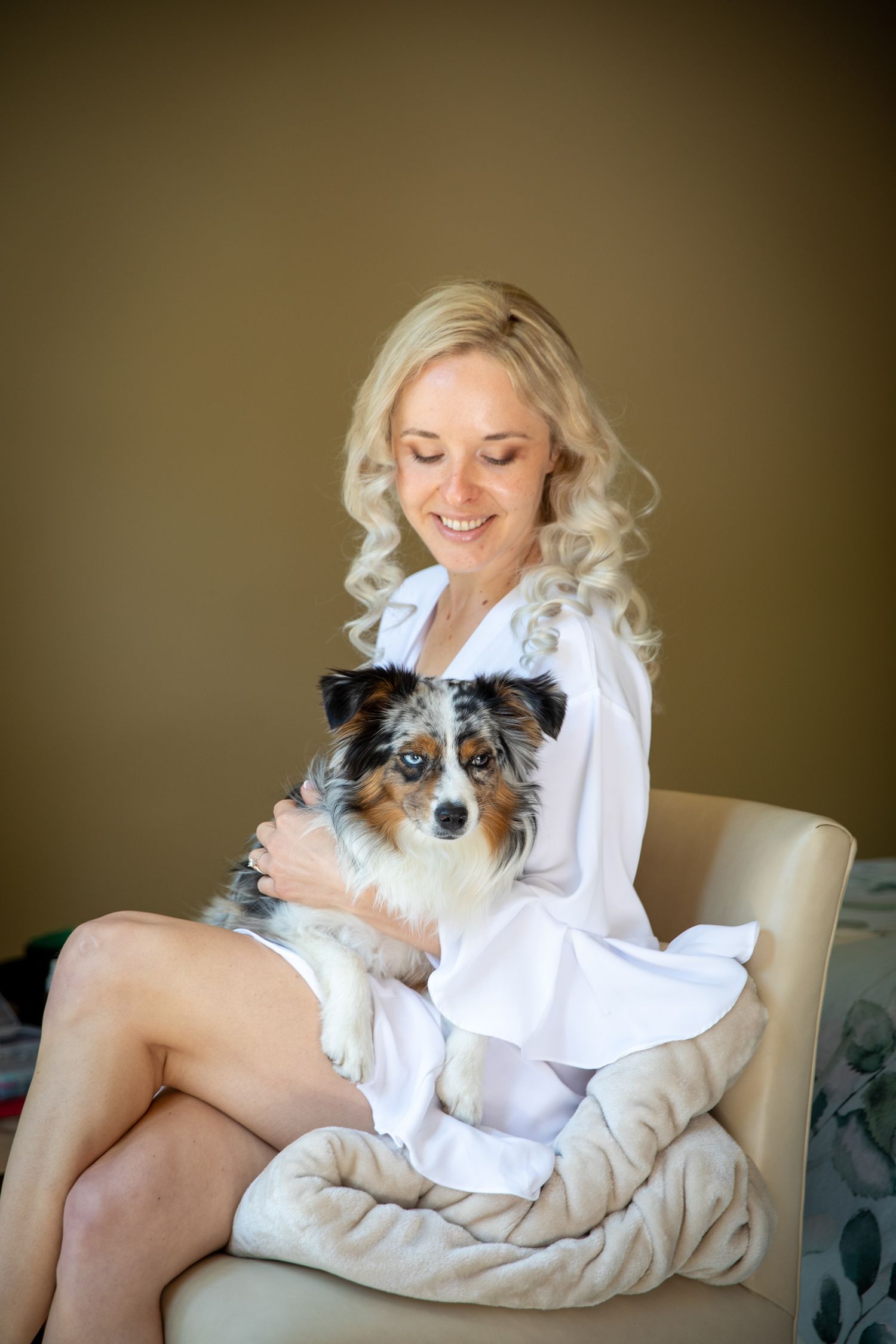 Smiling blonde woman in white robe sitting in chair holding a small tri-color Australian Shepherd dog In Canmore
