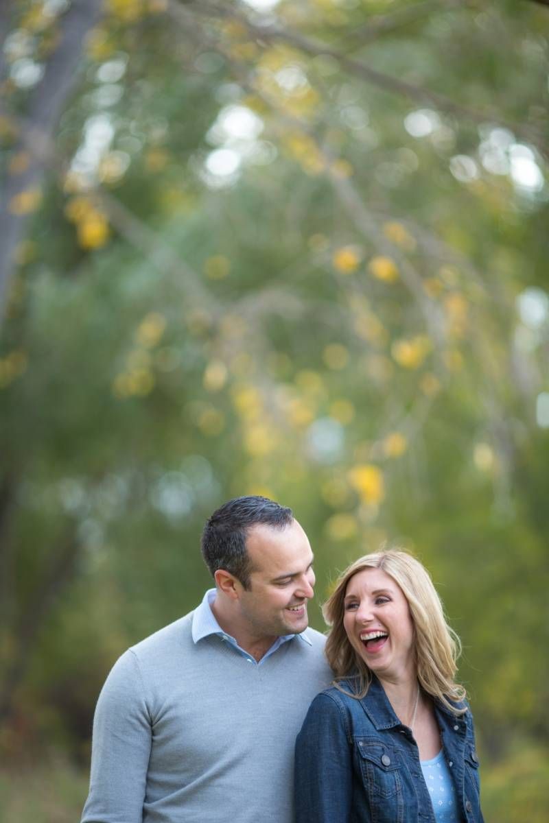 A laughing couple shares a joyful moment outdoors among autumn trees with yellow and green foliage in the background at Fish Creek Park.
