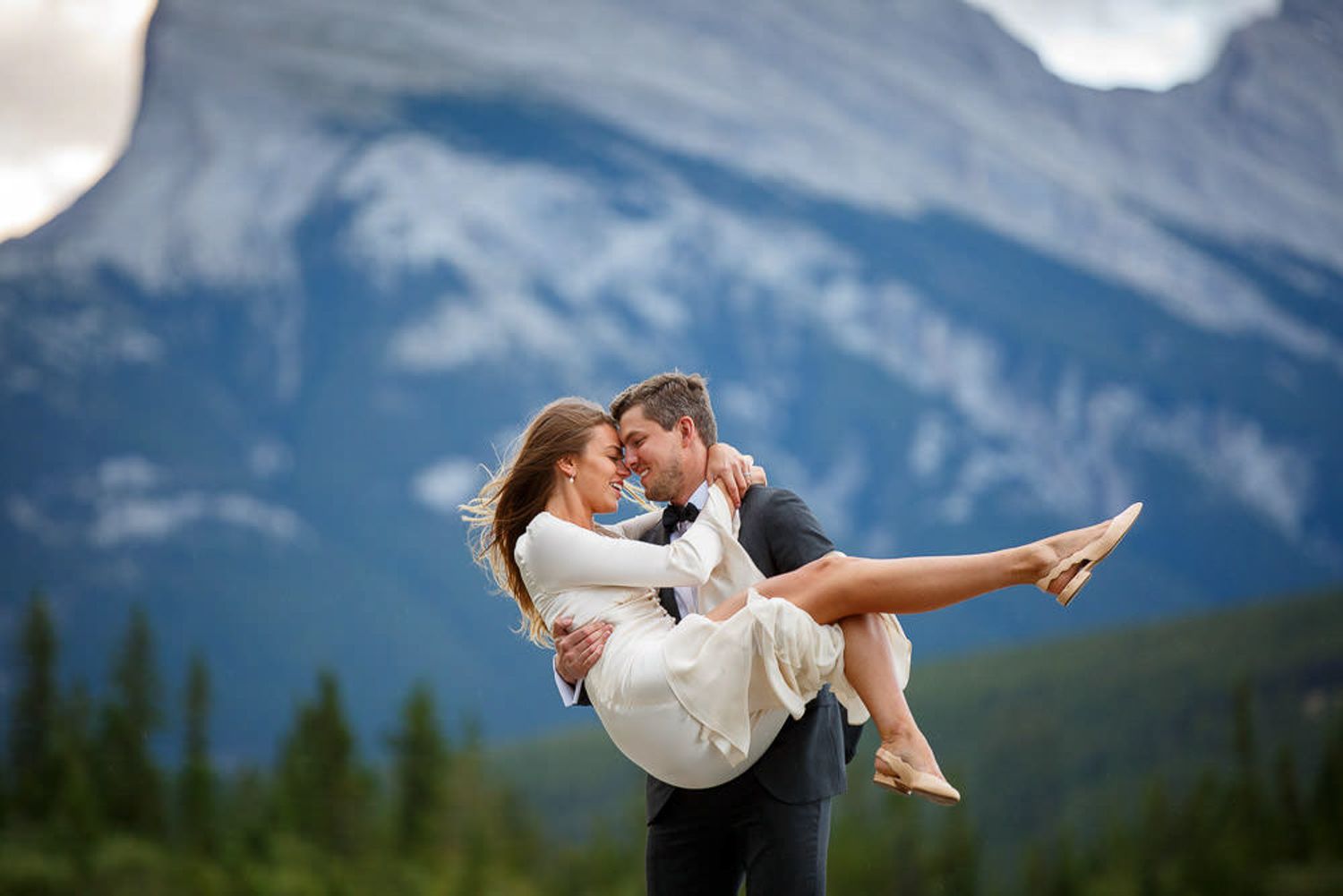 A couple in wedding attire shares an intimate moment against a backdrop of majestic mount Rundle peaks in background in Banff, Alberta.