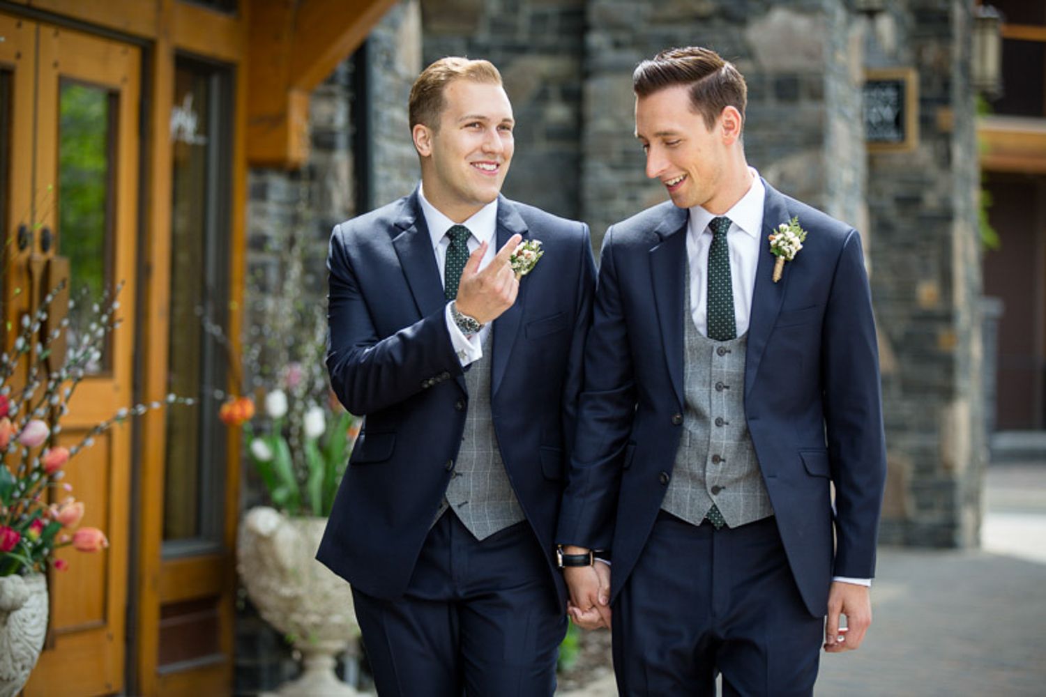 Two LGBTQ groomsmen in navy suits and gray vests share a candid moment while walking outside a stone building of Malcom Hotel in Canmore AB.