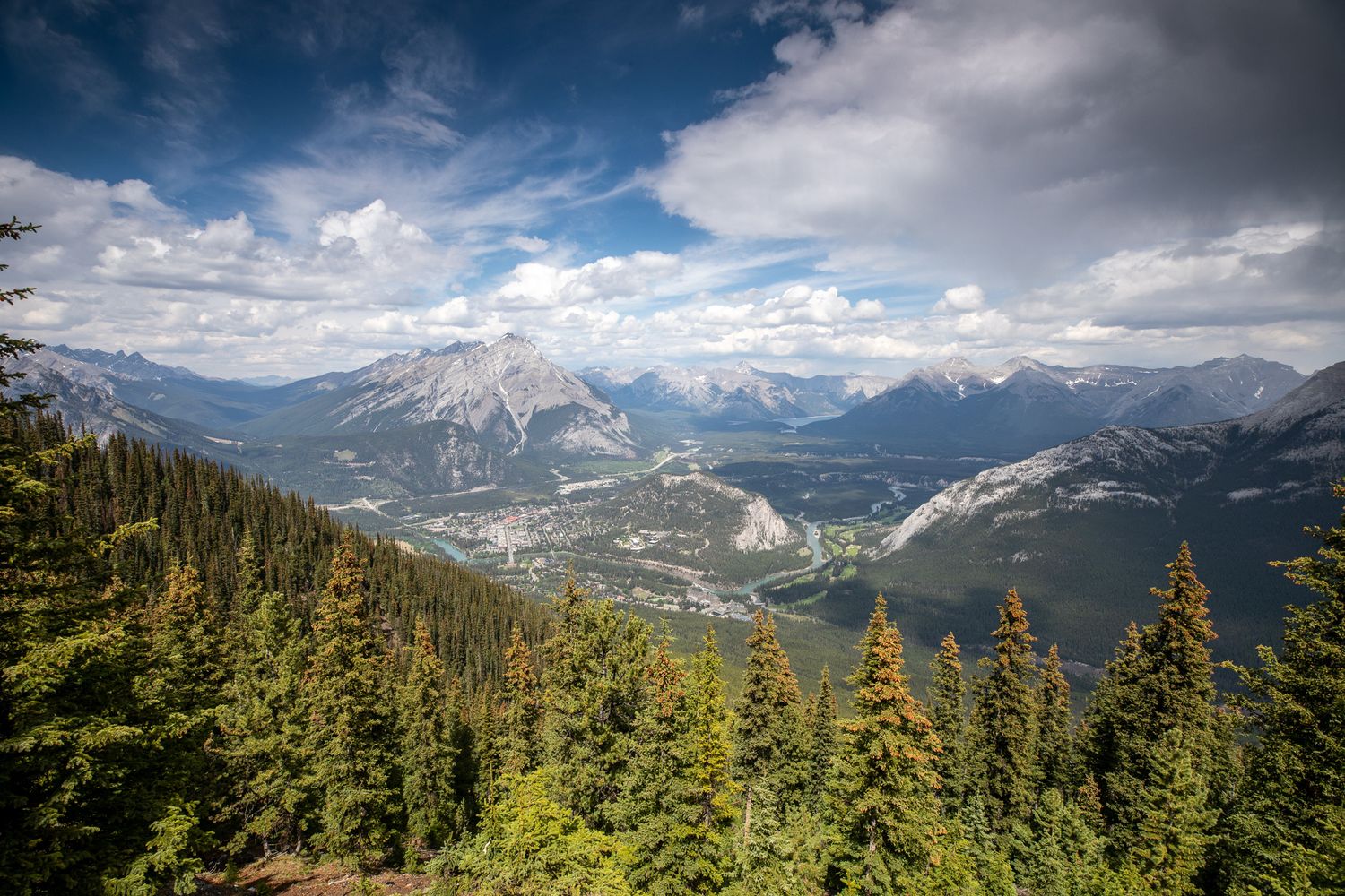 A panoramic mountain landscape with snow-capped peaks, evergreen forests, and cloudy skies from Mount Sulphur in Banff National Park, AB.