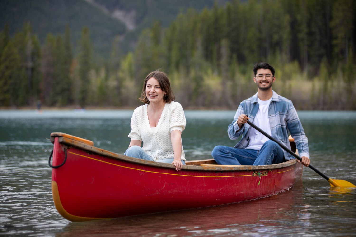 Two people enjoying a peaceful canoe ride in a red boat on a mountain lake surrounded by evergreen forest. Canmore Nordic Centre.