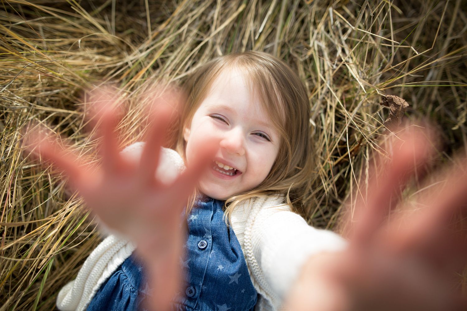 A playful sequence of photos shows a child in a blue shirt lying in hay while reaching towards the camera at Quarry Lake Park, Canmore, AB.