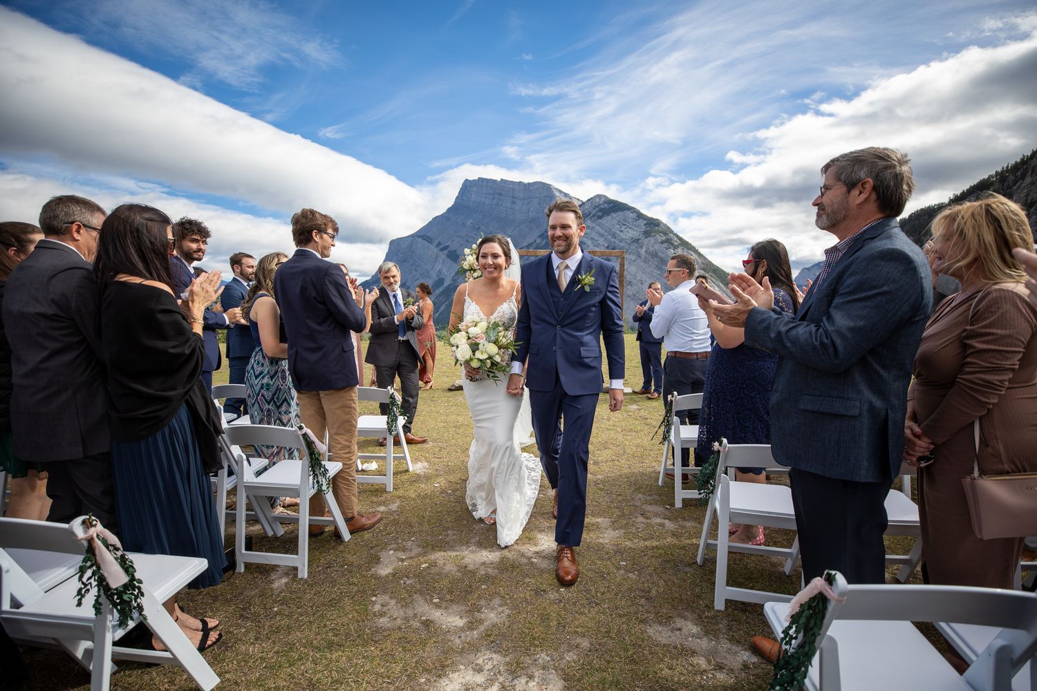 A scenic outdoor wedding ceremony takes place against a backdrop of majestic mountains and blue skies. Tunnel Mountain Reservoir Banff, AB