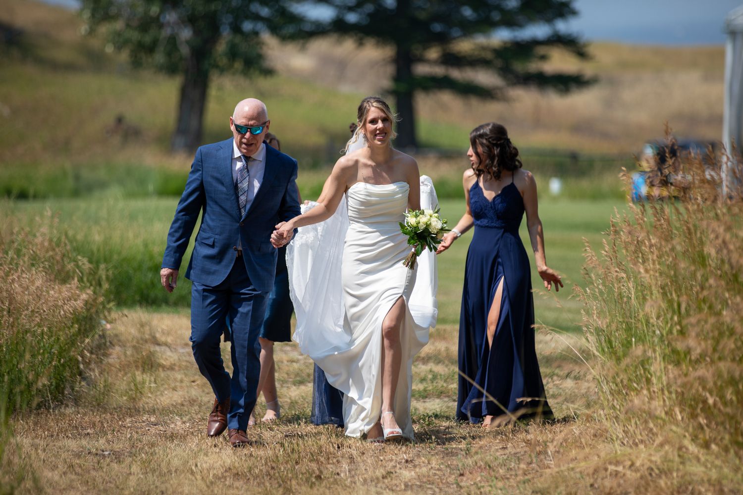 Bride in white gown walks with groom in blue suit and bridesmaid in navy dress outdoors on sunny wedding day at Swansea Ranch in Invermere