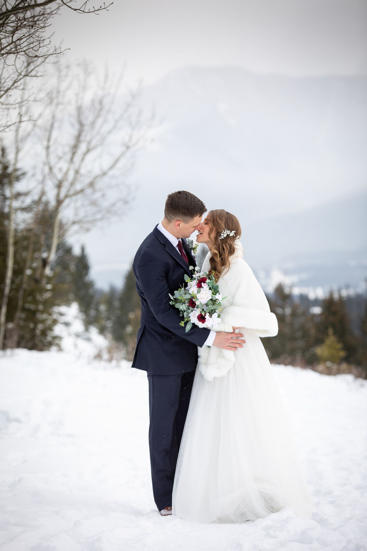 A romantic winter wedding photo with a couple embracing in the snow with mountain views and evergreen trees in background on Benchland trail