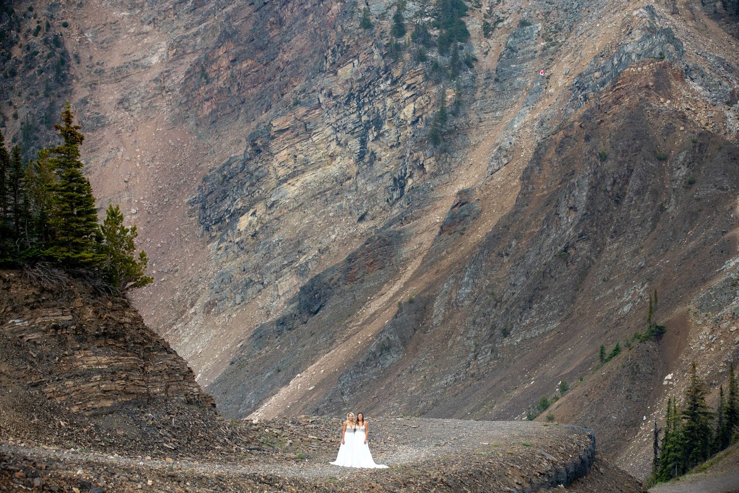 Couple in wedding attire embracing on a rocky mountain trail surrounded by dramatic cliffs and evergreen trees at Kicking Horse Resort, BC