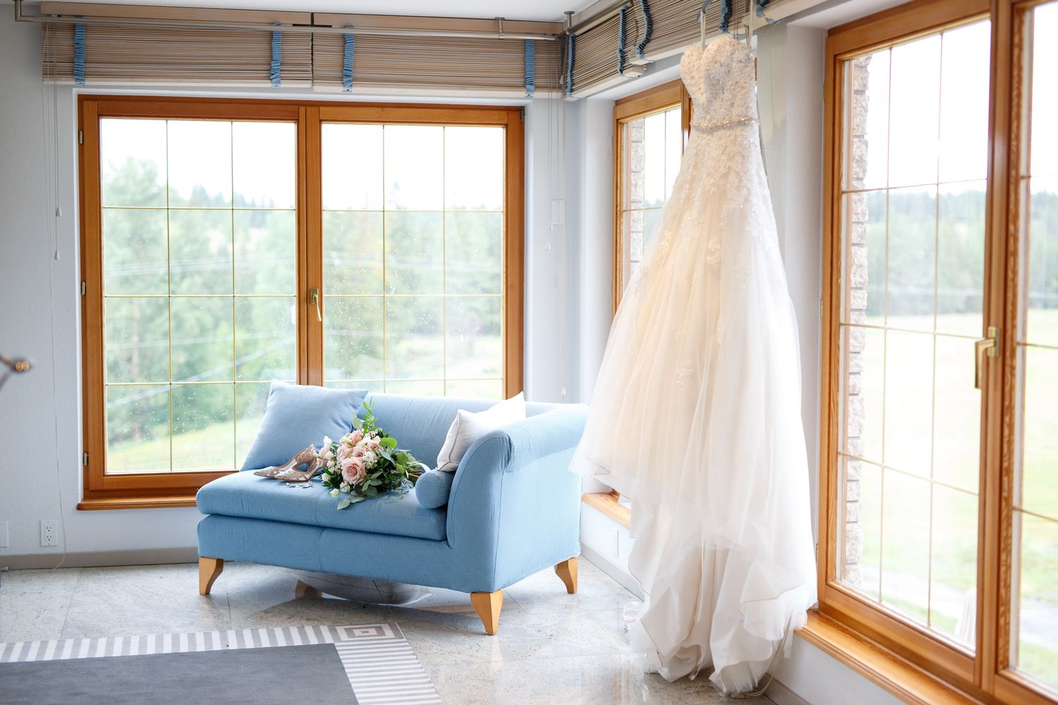White wedding gown hanging by sunlit windows beside a blue sofa with a bridal bouquet in an elegant bright room at Cherry creek estate