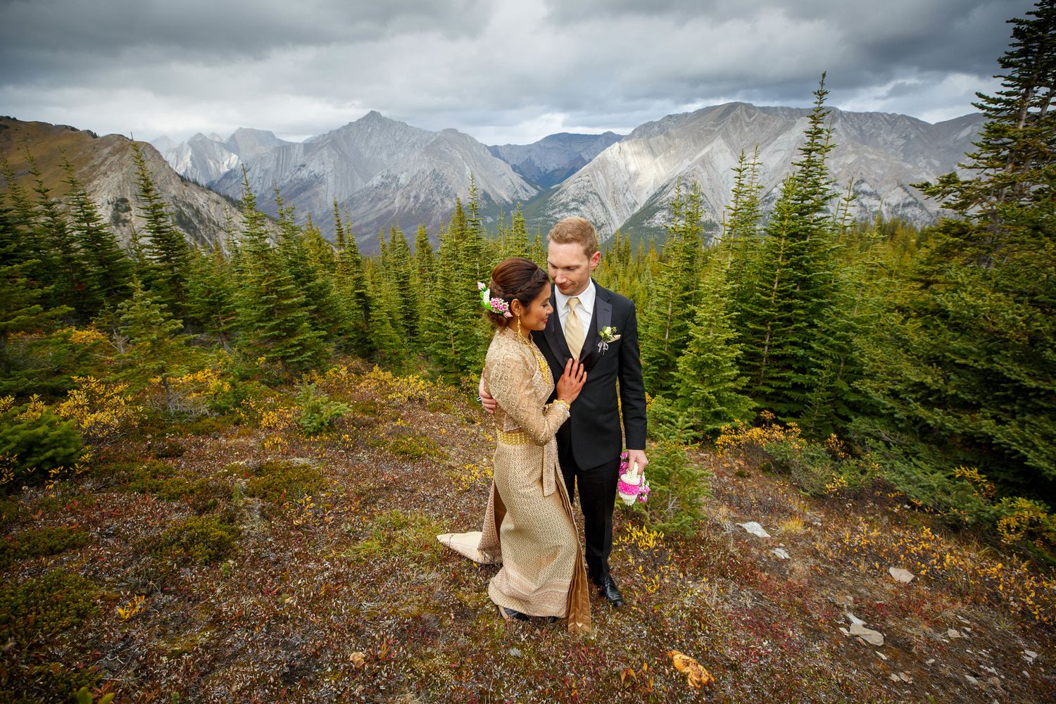 A couple in wedding attire embraces on a mountain ridge with evergreen trees and snow-capped peaks in the background in Banff, AB.
