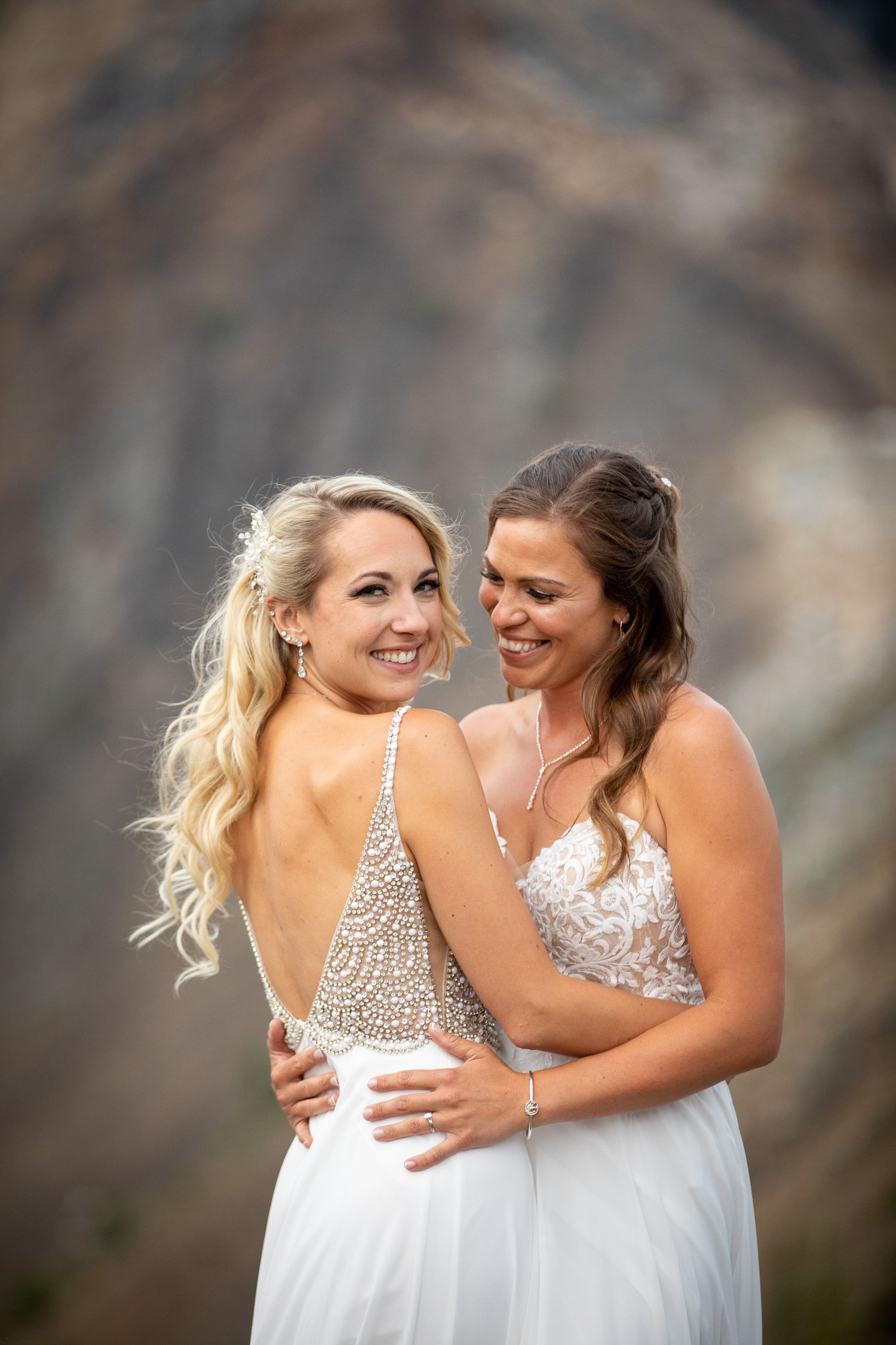 Two brides in white wedding dresses share an intimate embrace against a mountain backdrop during sunset at Kicking Horse resort in Golden