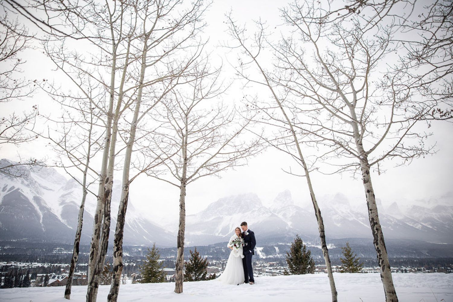A winter wedding photo session among bare aspen trees with snow-capped mountains in the background. Benchland trail Canmore, AB