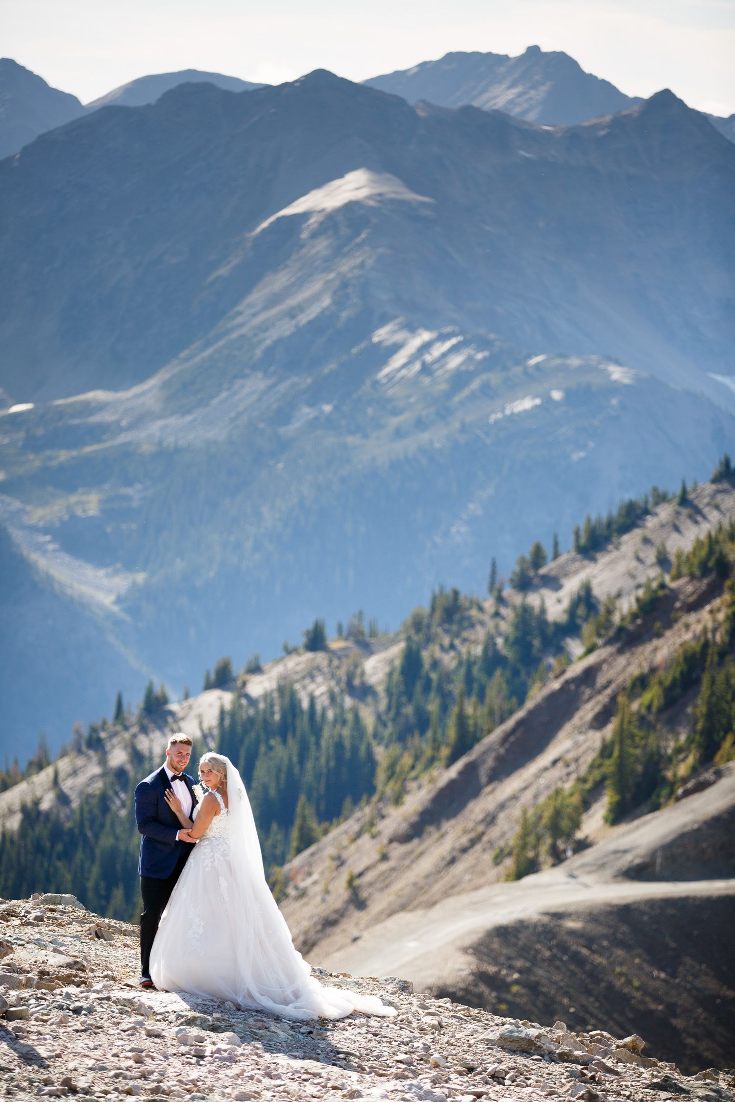 A couple in formal wedding attire poses atop a scenic mountain ridge at Kicking Horse resort with dramatic  peaks in the background. Golden.