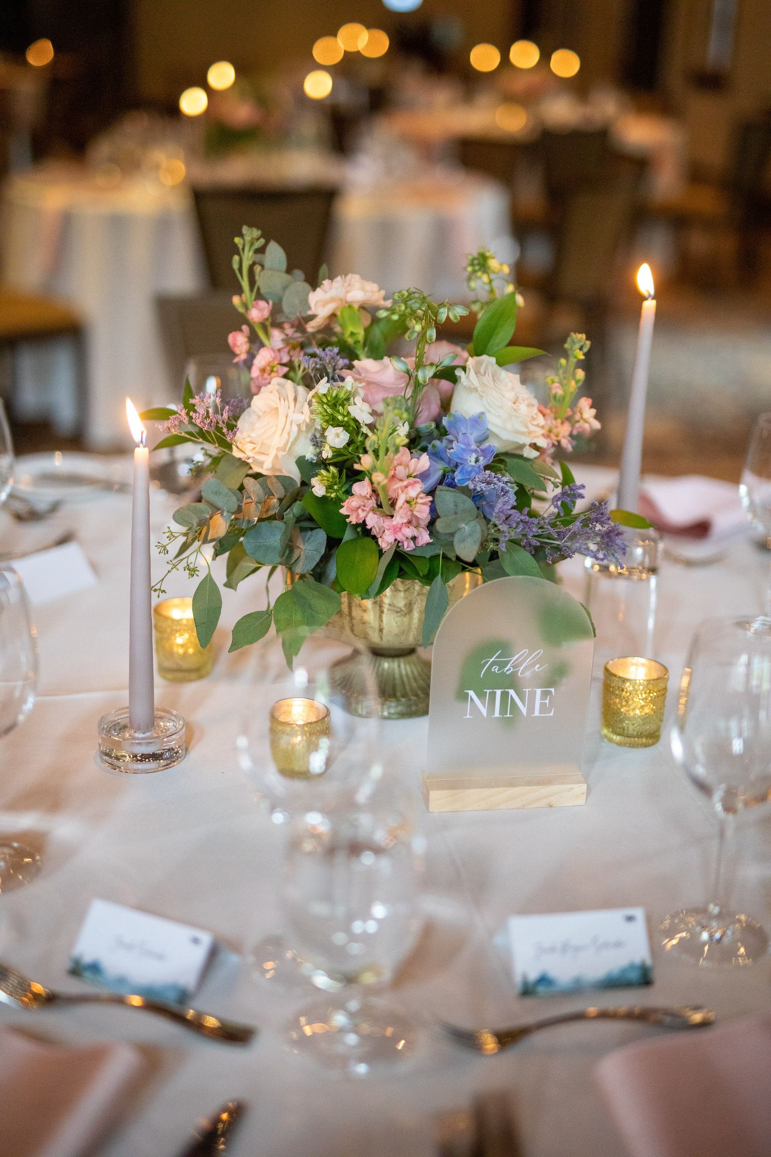Elegant wedding reception table nine with pastel floral center piece, taper candles, gold votives, and place cards on white linen Silvertip