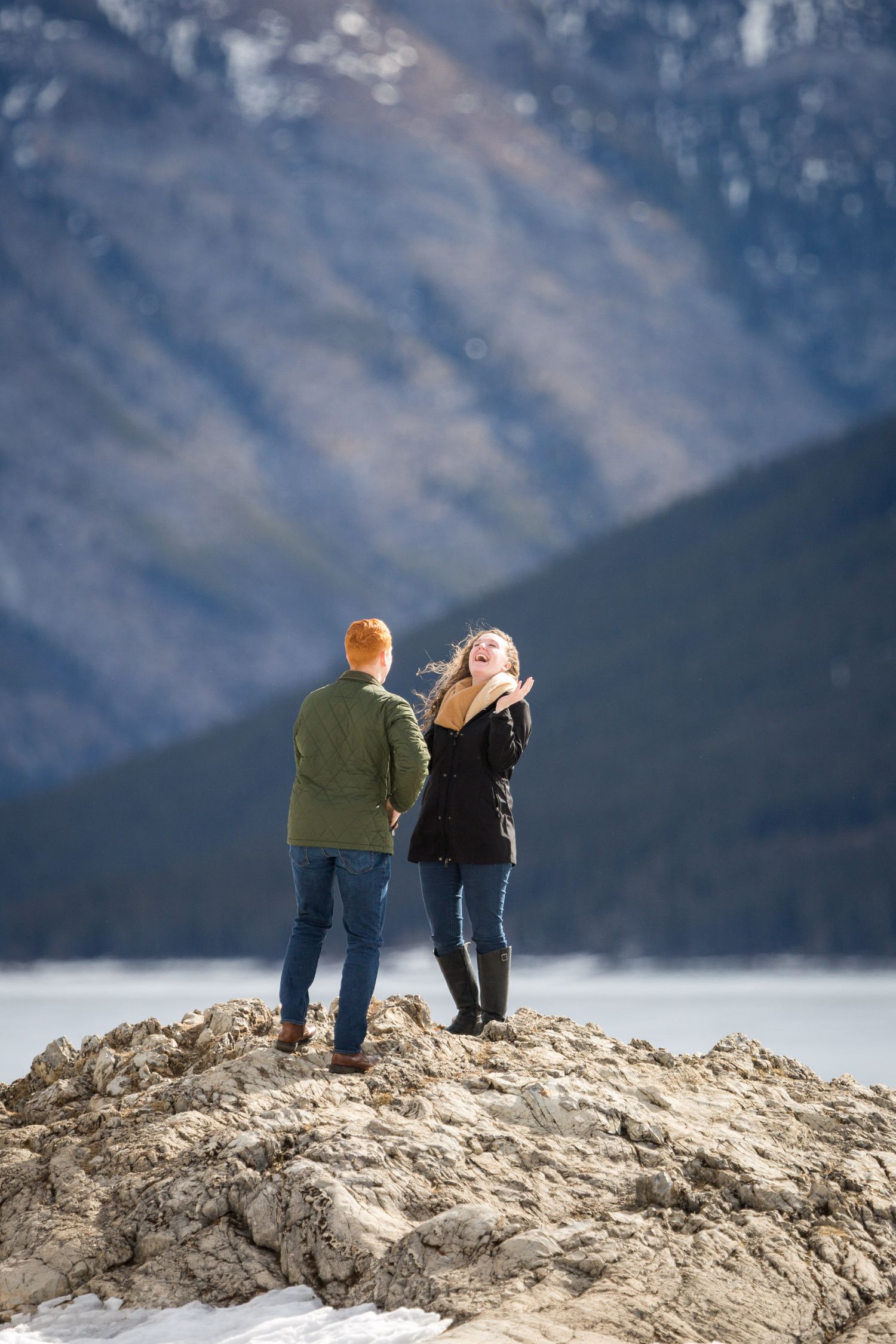 Couple standing on rocky outcrop at a surprise proposal with mountain lake backdrop in winterat Lake Minnewanka in Banff National Park.