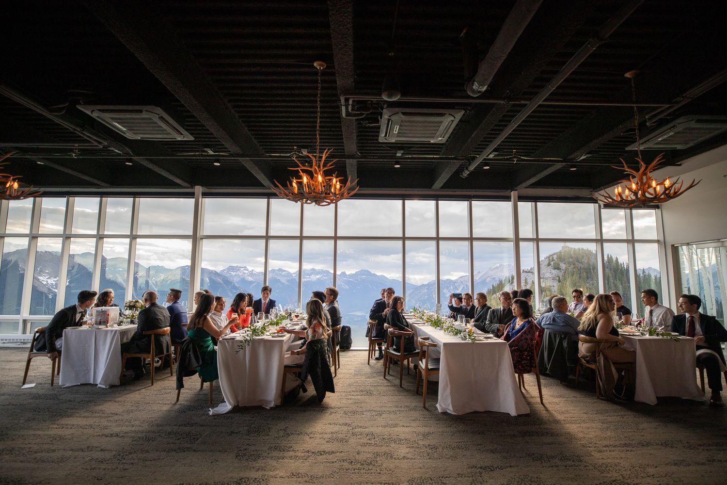 Elegant indoor dining event with mountain views through floor-to-ceiling windows, guests seated at long decorated tables.