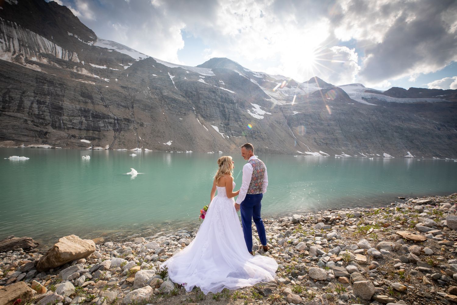 Bride and groom stand before turquoise glacial lake and snow-capped mountains in a stunning outdoor wedding photo. Lake-of-Hanging-Glacier.