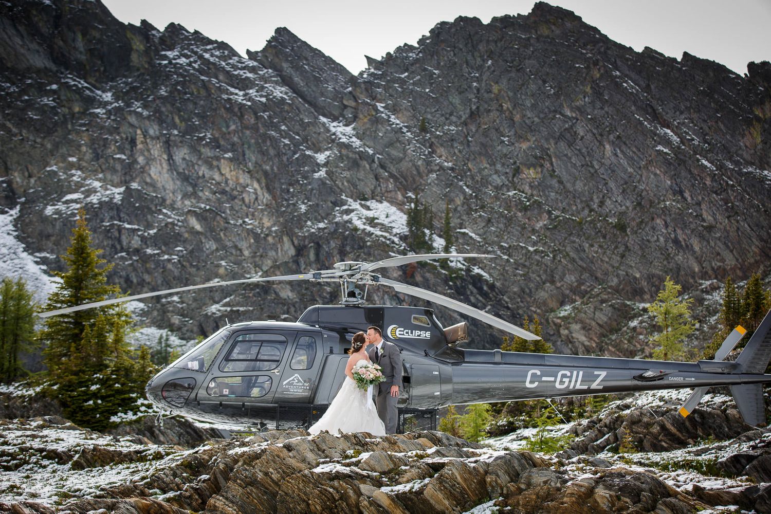 A bride and groom pose in front of a grey helicopter in the backcountry of Kimberley, British Columbia.