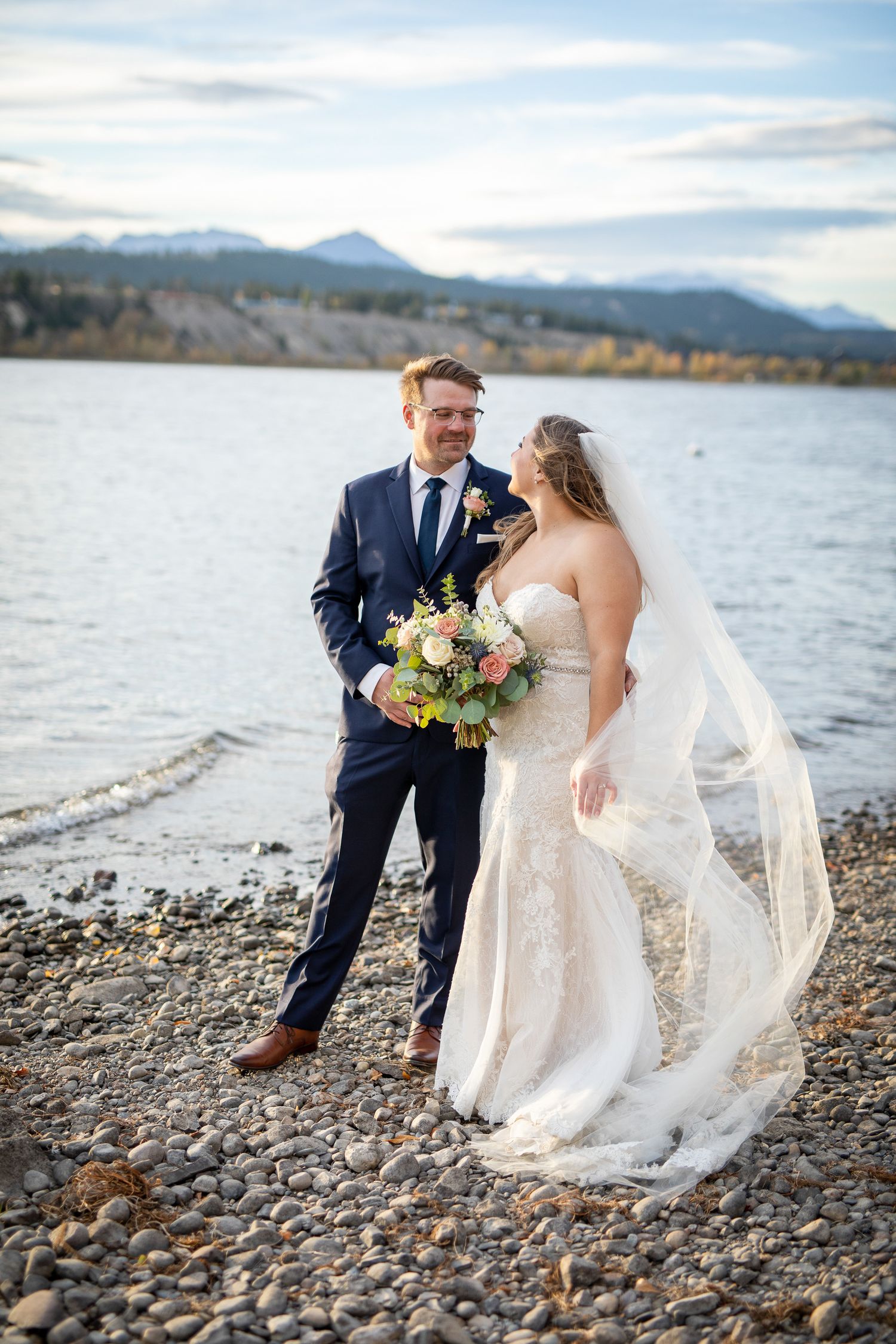 A romantic wedding couple poses on a rocky Windermere lakeshore with mountains in the background at sunset. Invermere, British Columbia.
