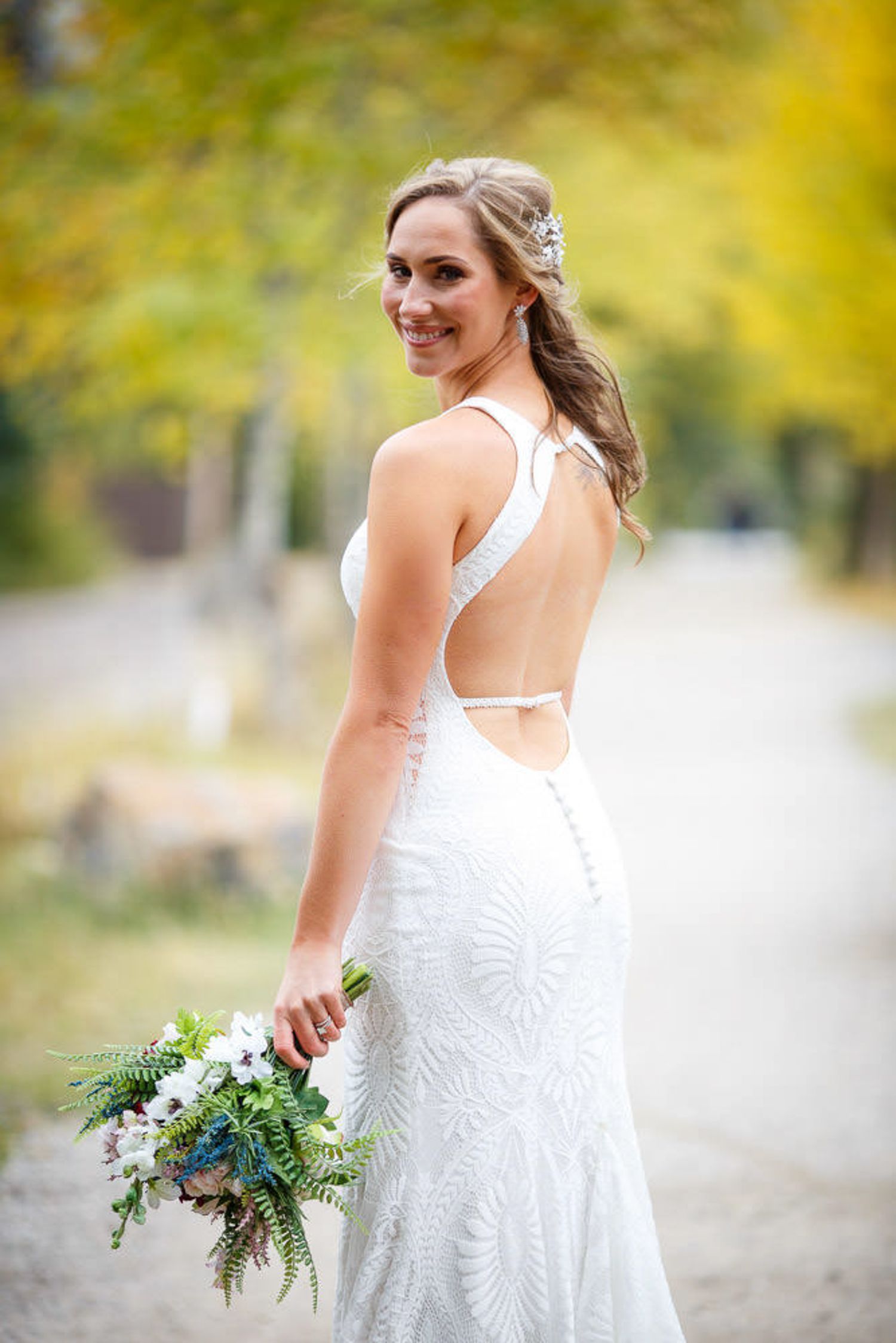 A bride in a backless white lace wedding dress holds a rustic bouquet of flowers and greenery in an outdoor setting of Canmore Alberta.