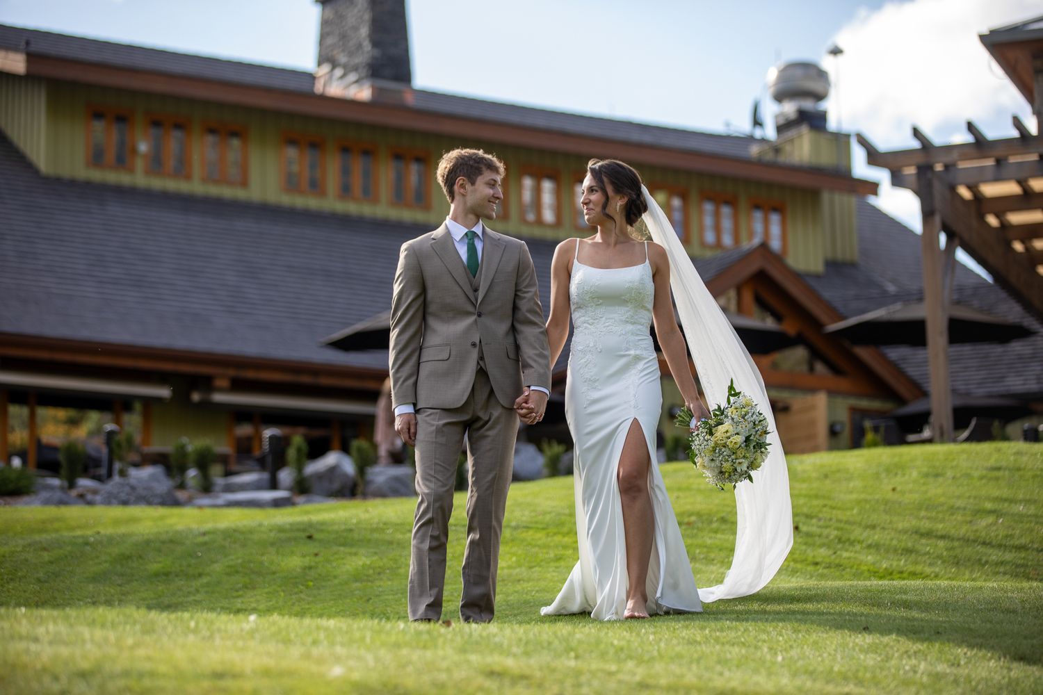 A couple in formal wedding attire walks hand in hand across a green lawn at the venue Silvertip resort in Canmore, Alberta.
