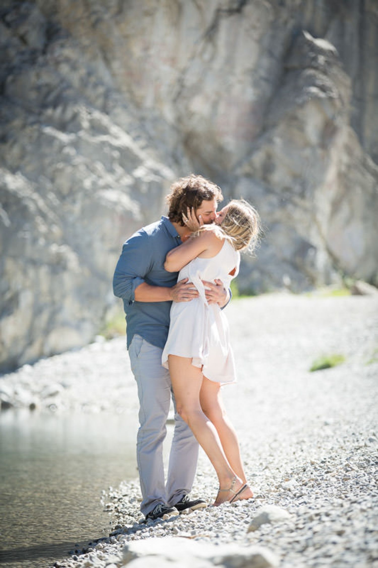 A romantic couple embraces on a rocky beach shore with cliffs and water in the background Canmore reservoir.
