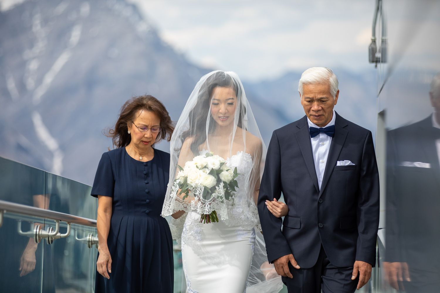 Bride in white gown and veil walks with parents toward mountain backdrop at outdoor wedding ceremony.